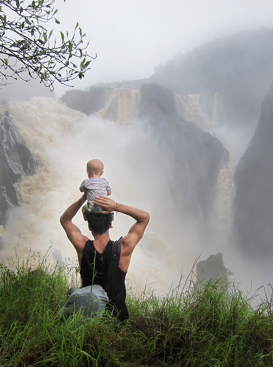 Man with a baby balanced on his head, overlooking a waterfall in flood